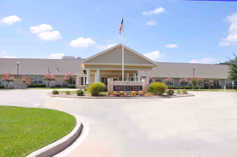 Front exterior view of a single-story senior living facility building with a covered entrance, an American flag on a flagpole, and landscaping including bushes and flowering trees under a blue sky with some clouds.