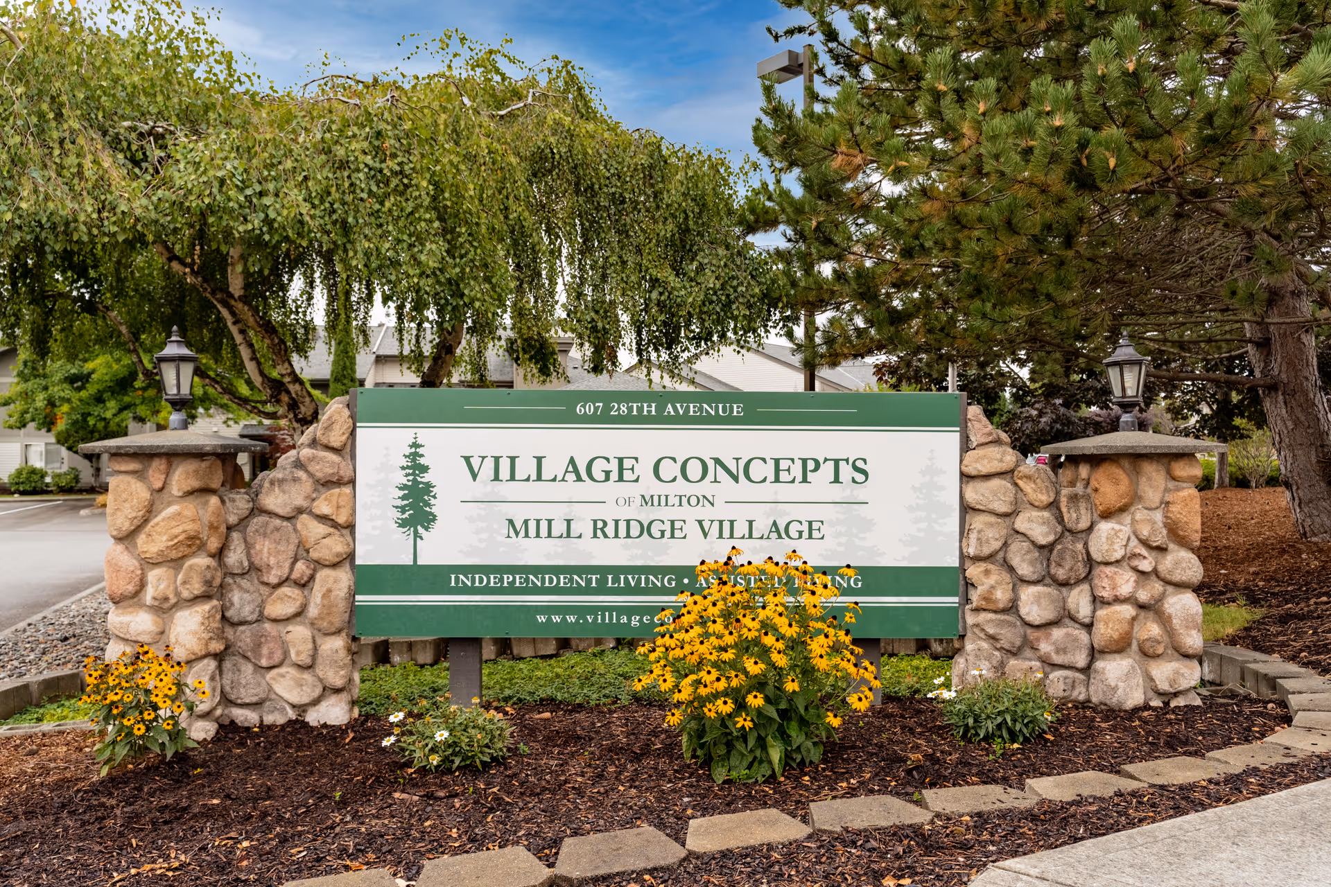 Stone-pillared roadside sign for Village Concepts Mill Ridge Village surrounded by flowers and trees.