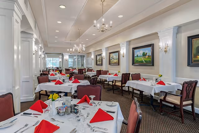 A spacious dining room with multiple tables covered in white tablecloths, each set with red folded napkins, glassware, and silverware. The room features elegant chandeliers, wall sconces, framed landscape paintings, and large windows letting in natural light. Chairs with red upholstery surround the tables.