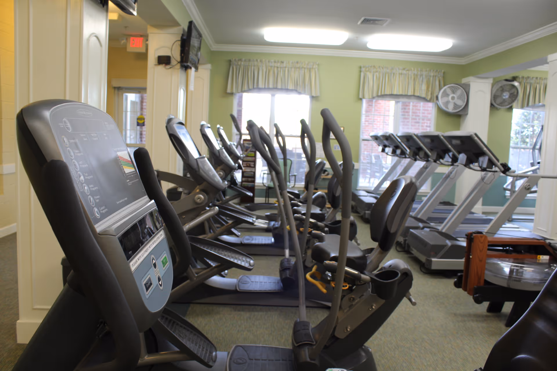 Interior view of a fitness room with multiple exercise machines including treadmills, elliptical trainers, and stationary bikes. The room has large windows with curtains, wall-mounted fans, and a television on the wall.