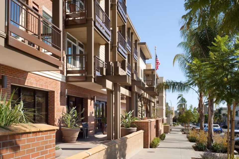 Exterior view of a multi-story residential building with balconies, brick and beige siding, potted plants, and a sidewalk lined with palm trees and other greenery under a clear blue sky.