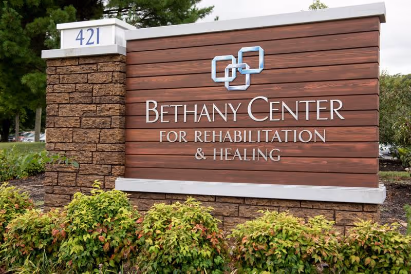 A large outdoor sign for Bethany Center for Rehabilitation & Healing made of wood and stone with the address number 421 on the left side, surrounded by green bushes and trees.