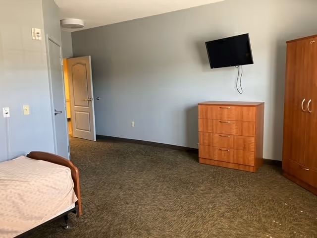Furnished senior bedroom with a bed, wooden dresser and wardrobe, a wall-mounted TV, and an open doorway.
