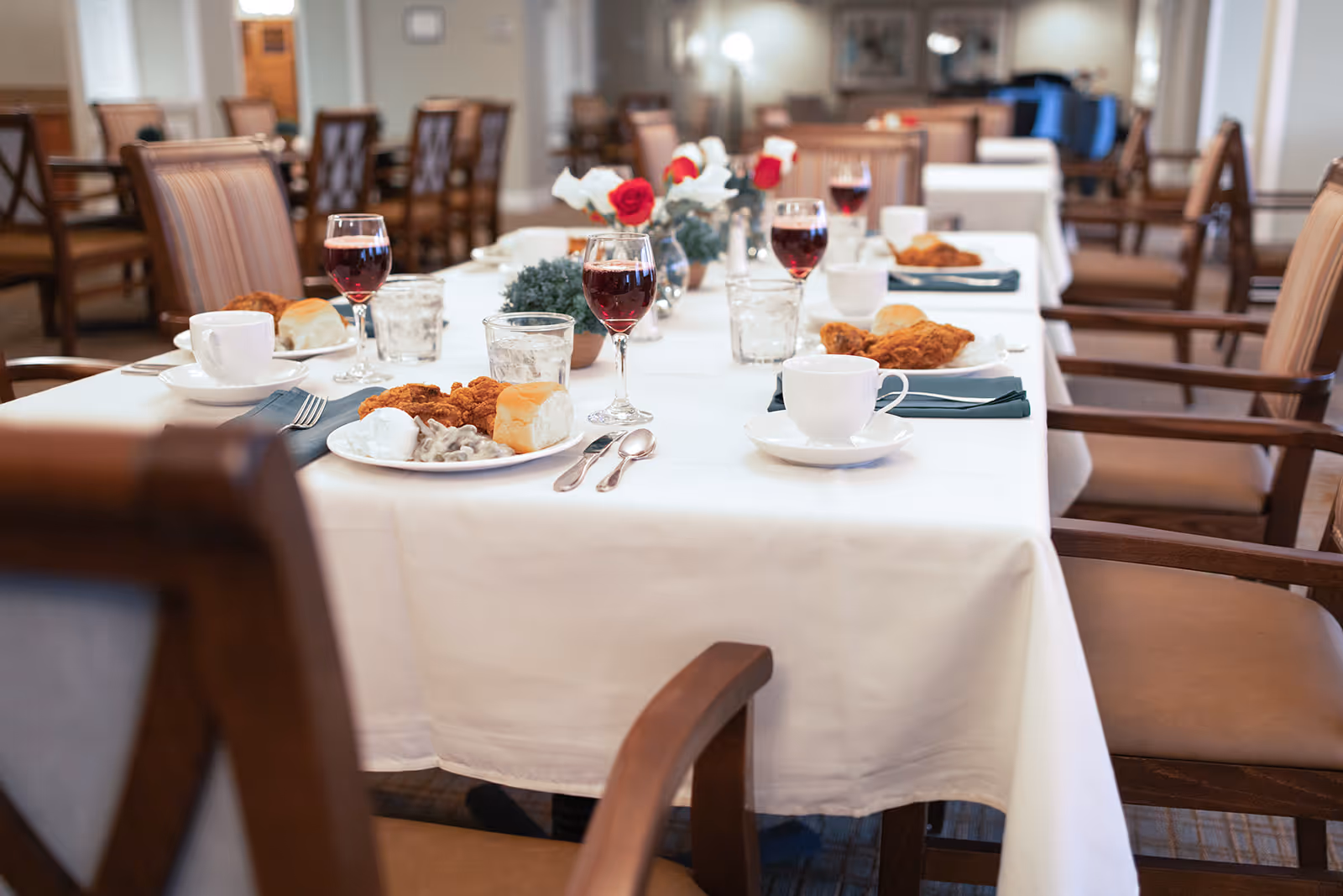 A communal dining room table set with plates of fried chicken, rolls, wine glasses and coffee cups on a white tablecloth.