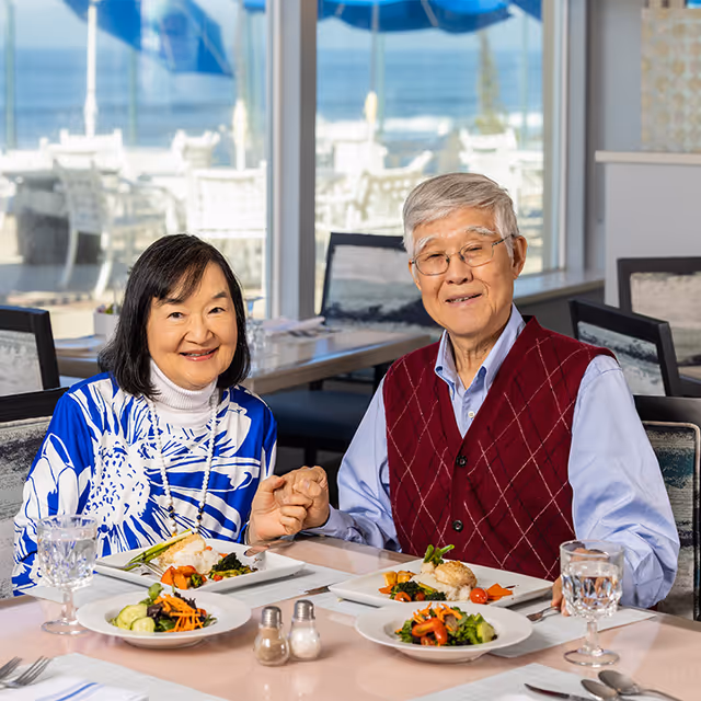 An elderly couple sitting at a dining table in a bright room with large windows showing an outdoor view. They are holding hands and smiling at the camera. Plates of food and glasses of water are on the table in front of them.