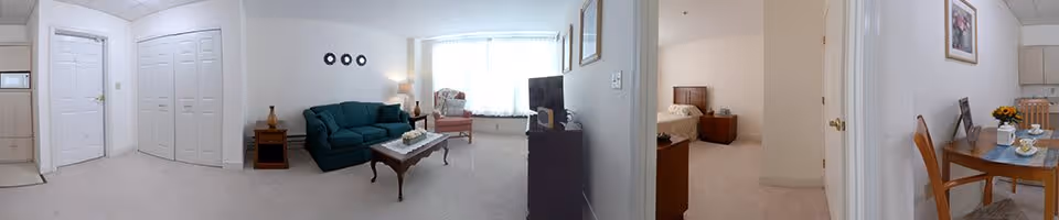 Panoramic view of an assisted living residence interior showing a living room with a green sofa, coffee table, armchair, and TV stand. Adjacent to the living room is a bedroom with a bed and nightstand, and a small dining area with a table and chairs. The walls are light-colored, and the space is well-lit with natural light from a large window.