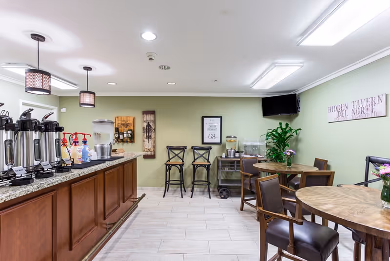 A cozy dining area with wooden tables and chairs, a counter with coffee dispensers and condiments, green walls decorated with framed art and a sign reading 'Hidden Tavern Del Norte', and a small TV mounted in the corner.