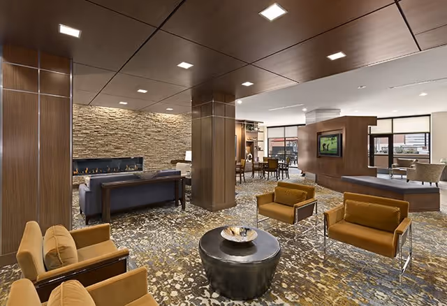 A spacious and modern senior living facility common area featuring comfortable seating with mustard-colored armchairs, a round black coffee table, a stone accent wall with a built-in fireplace, and a flat-screen TV mounted on a wooden partition. The room has a patterned carpet, wood-paneled columns and ceiling, and large windows letting in natural light.