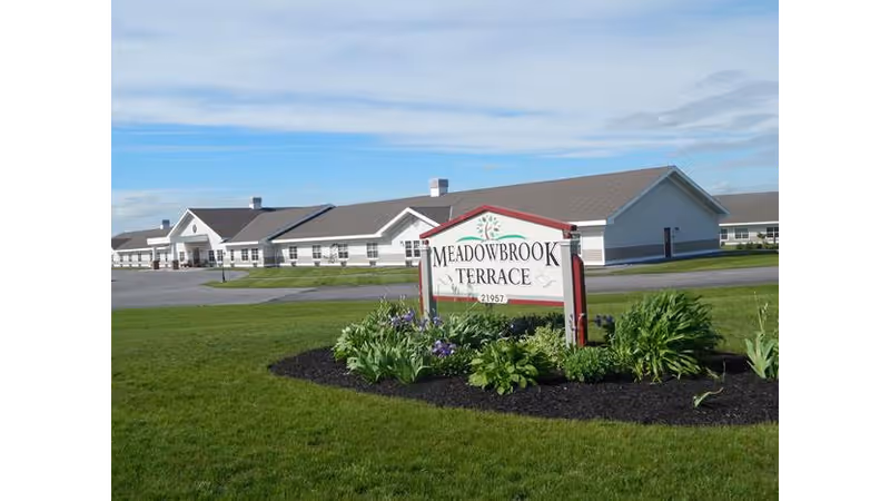 Exterior view of Meadowbrook Terrace Assisted Living Facility showing a long, single-story building with a gray roof and white walls. In the foreground, there is a landscaped area with green grass, plants, and flowers surrounding a sign that reads 'Meadowbrook Terrace 21957'. The sky is partly cloudy with blue patches.