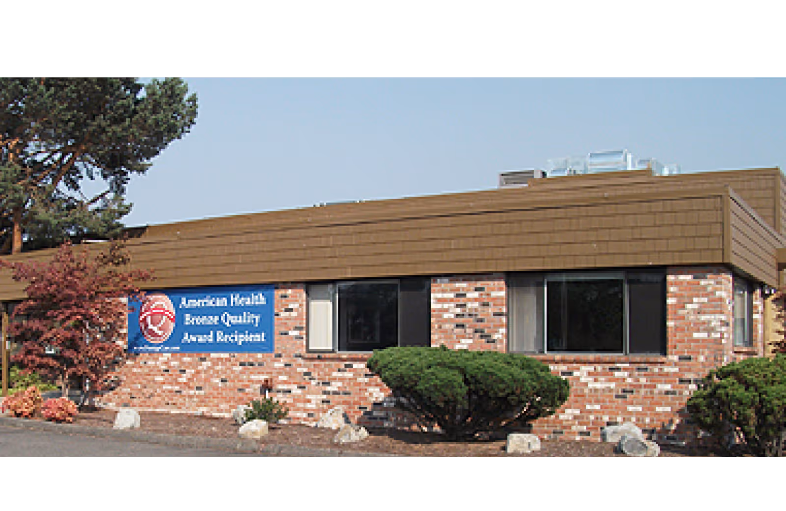 One-story brick building with a brown shingled roof, shrubs in front, and a blue award sign on the facade.