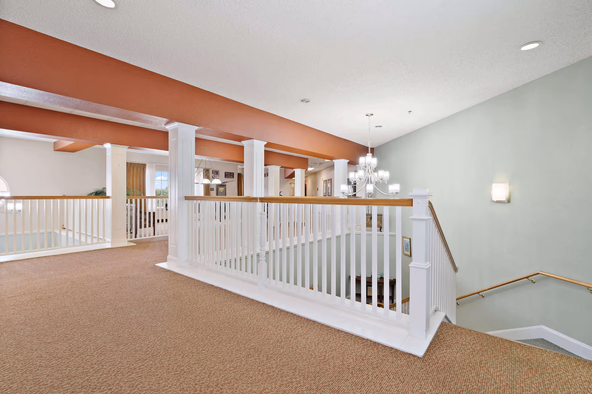 Second-floor interior common area with carpeted hallway, white railing and columns overlooking a stairwell and a chandelier.