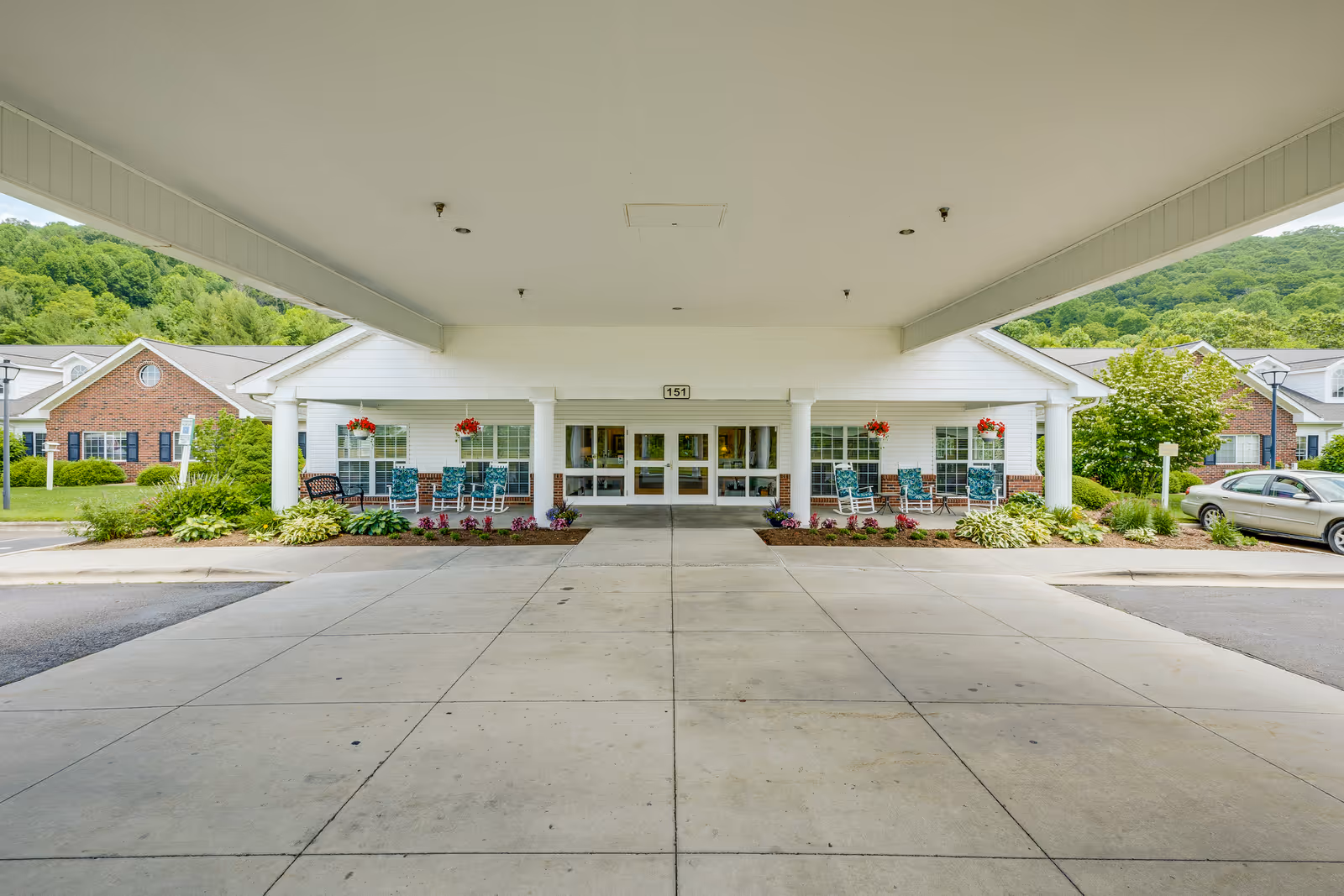 Covered porte-cochère and entrance to an assisted living building with rocking chairs, hanging flower baskets, and landscaped beds.