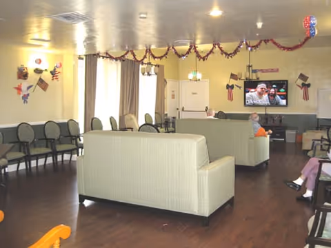 A common area in a retirement center with beige sofas arranged facing a wall-mounted television. The room has wooden flooring, several chairs lined up against the walls, and patriotic decorations including American flags and red, white, and blue garlands hanging from the ceiling. Two elderly people are seated on the sofas watching TV.