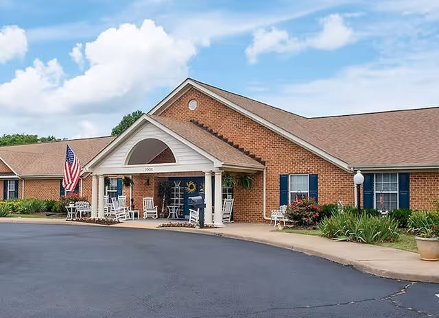 Exterior view of a single-story brick building with a covered entrance supported by white columns. The entrance area has white rocking chairs and an American flag displayed. There are shrubs and plants along the building, and the sky is partly cloudy.