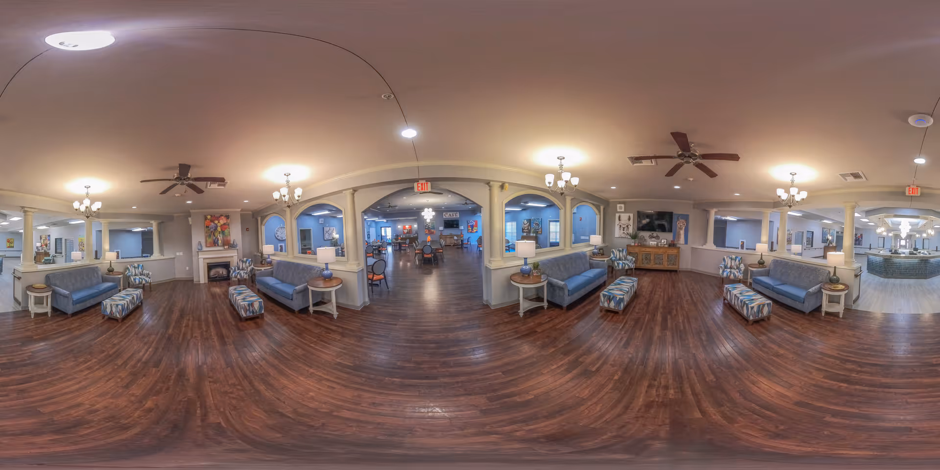 Panoramic interior lobby of a senior living facility with multiple sofas, ottomans, chandeliers, ceiling fans and wood floors opening into a dining area.