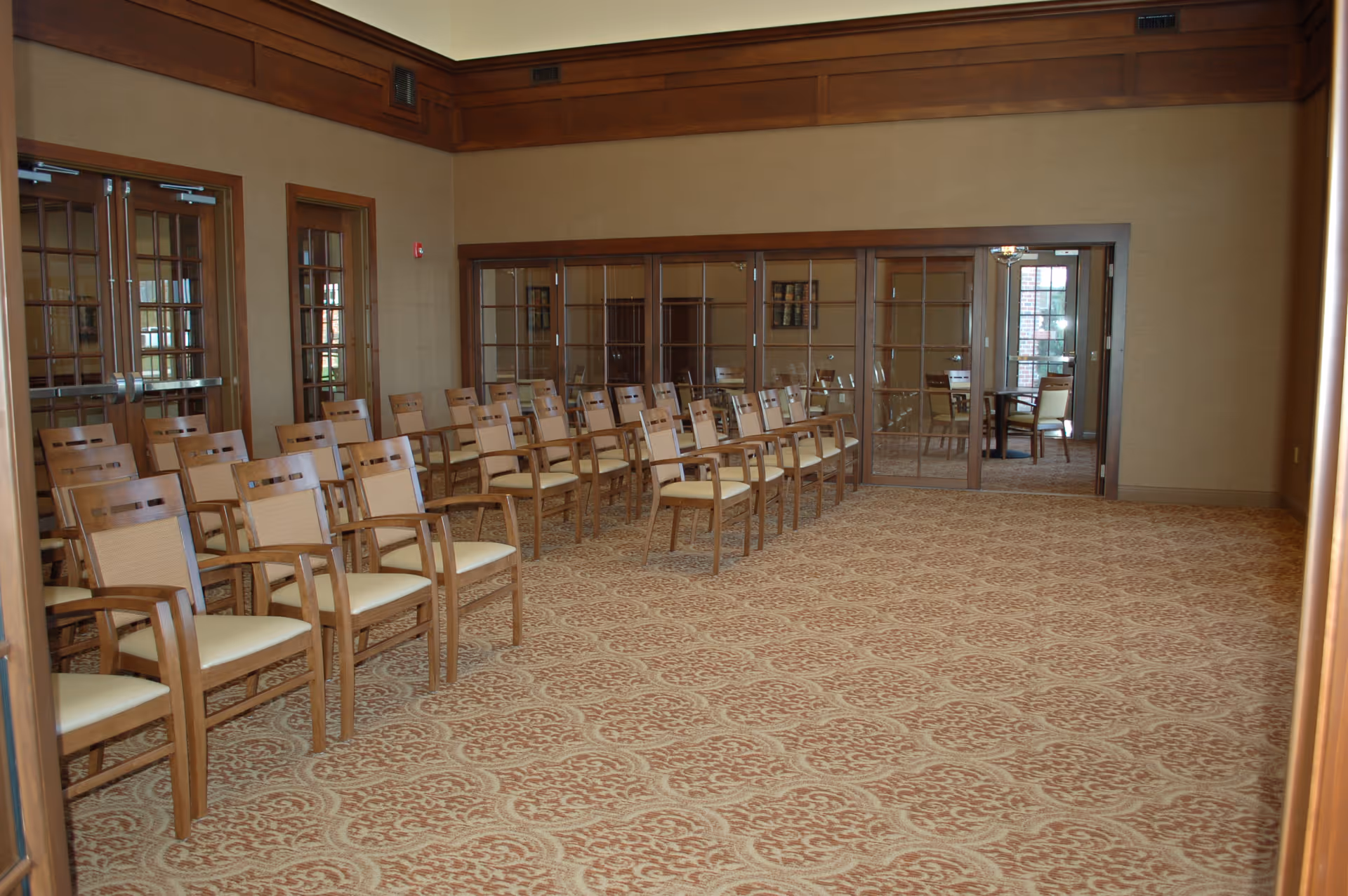 A spacious room with rows of wooden chairs with beige cushions arranged in a neat formation on a patterned carpet. The room has beige walls with wooden trim and large glass-paneled doors leading to another room with additional seating and natural light coming through windows.