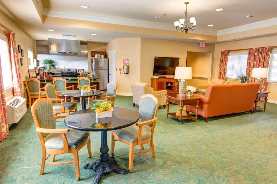 A bright and spacious common area in a senior living facility featuring a small kitchen with stainless steel appliances, several round tables with chairs, and a living room area with sofas, armchairs, a coffee table, side tables with lamps, and a flat-screen TV mounted on a wooden cabinet. The room has large windows with floral curtains and green patterned carpet.