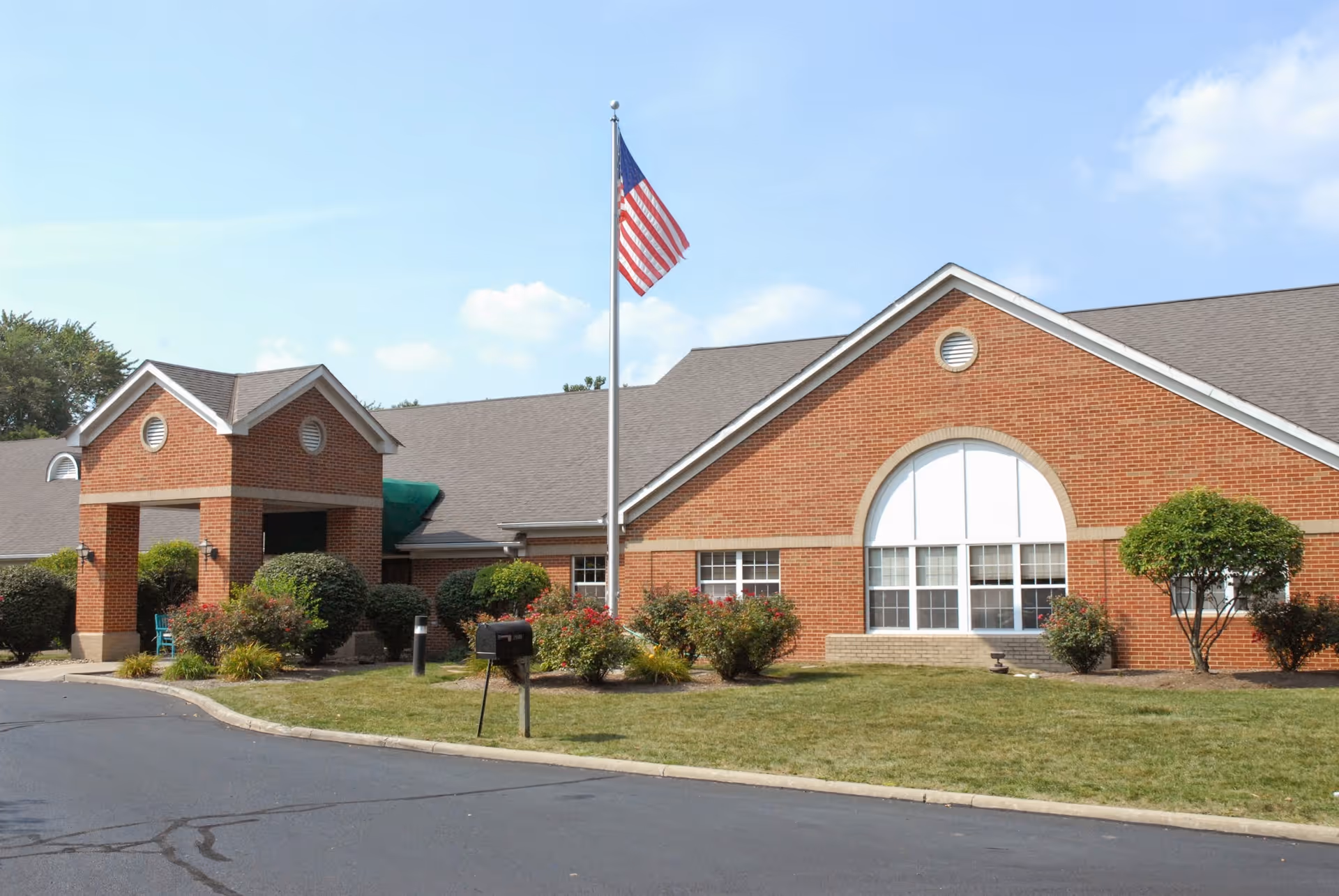 Exterior view of a single-story brick building with a peaked roof, featuring a covered entrance supported by brick columns. There is an American flag on a flagpole in front of the building, surrounded by neatly trimmed bushes and a lawn. The sky is clear with a few clouds.