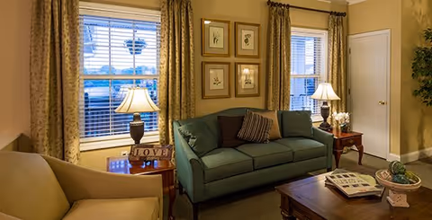 Cozy living room with a green sofa, armchair, coffee table, table lamps, and framed artwork between two windows.
