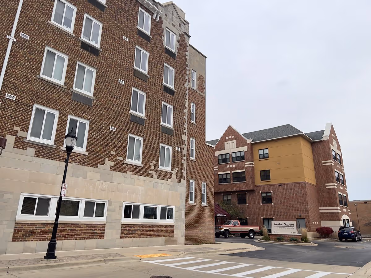 Exterior view of Avalon Square, a multi-story brick senior living building with an entrance, parked vehicles, and a streetlamp.