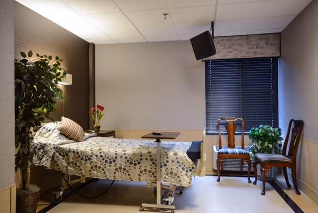 A furnished patient bedroom with a hospital-style bed, bedside table, chairs, a wall-mounted TV, and potted plants.