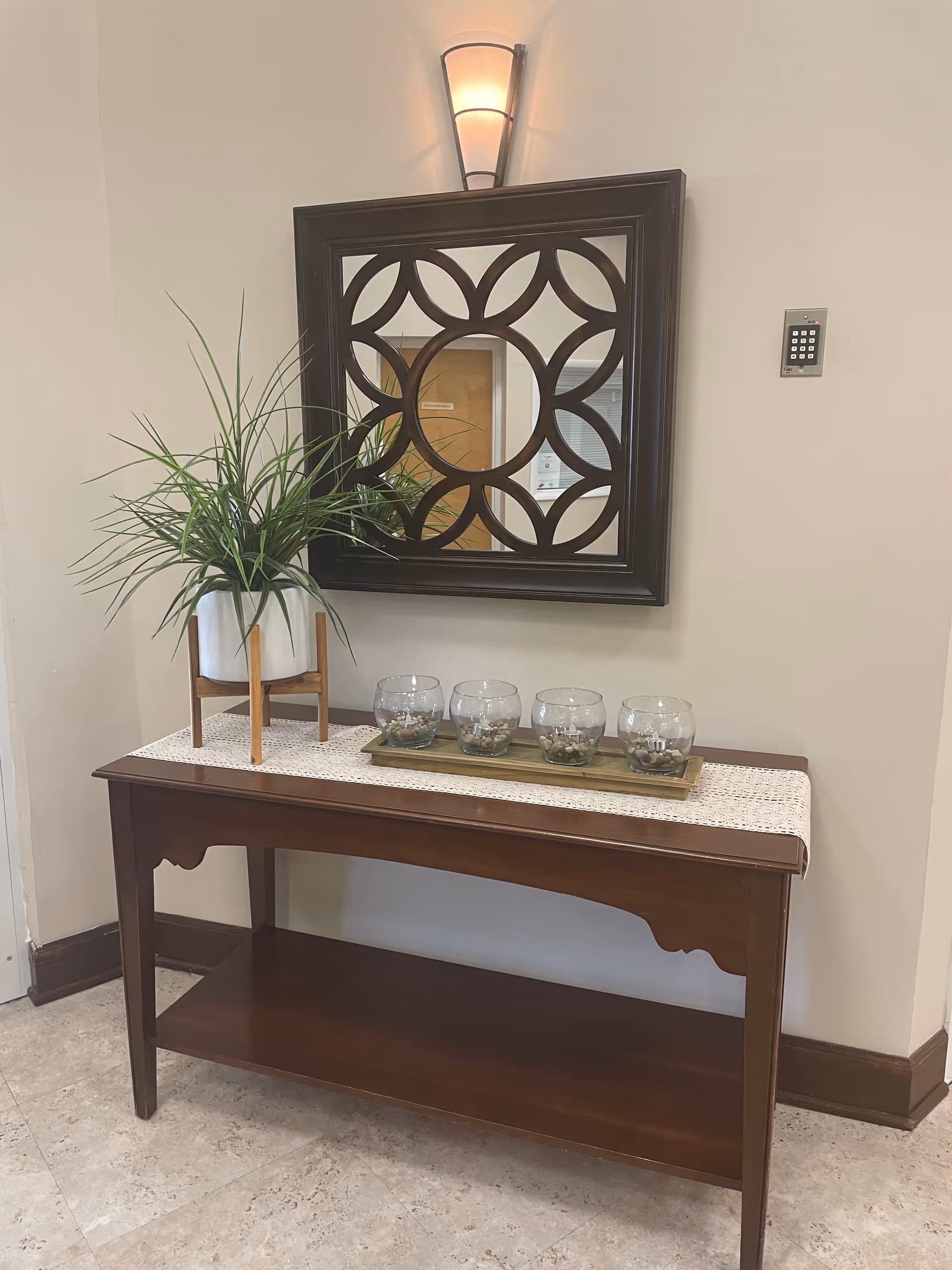 A wooden console table with a white runner, holding a potted green plant in a white pot on a wooden stand and four glass candle holders with pebbles inside on a rectangular tray. Above the table is a decorative square mirror with a dark wooden frame featuring a circular and petal-like pattern. A wall-mounted light fixture is positioned above the mirror, and a keypad is mounted on the wall to the right.
