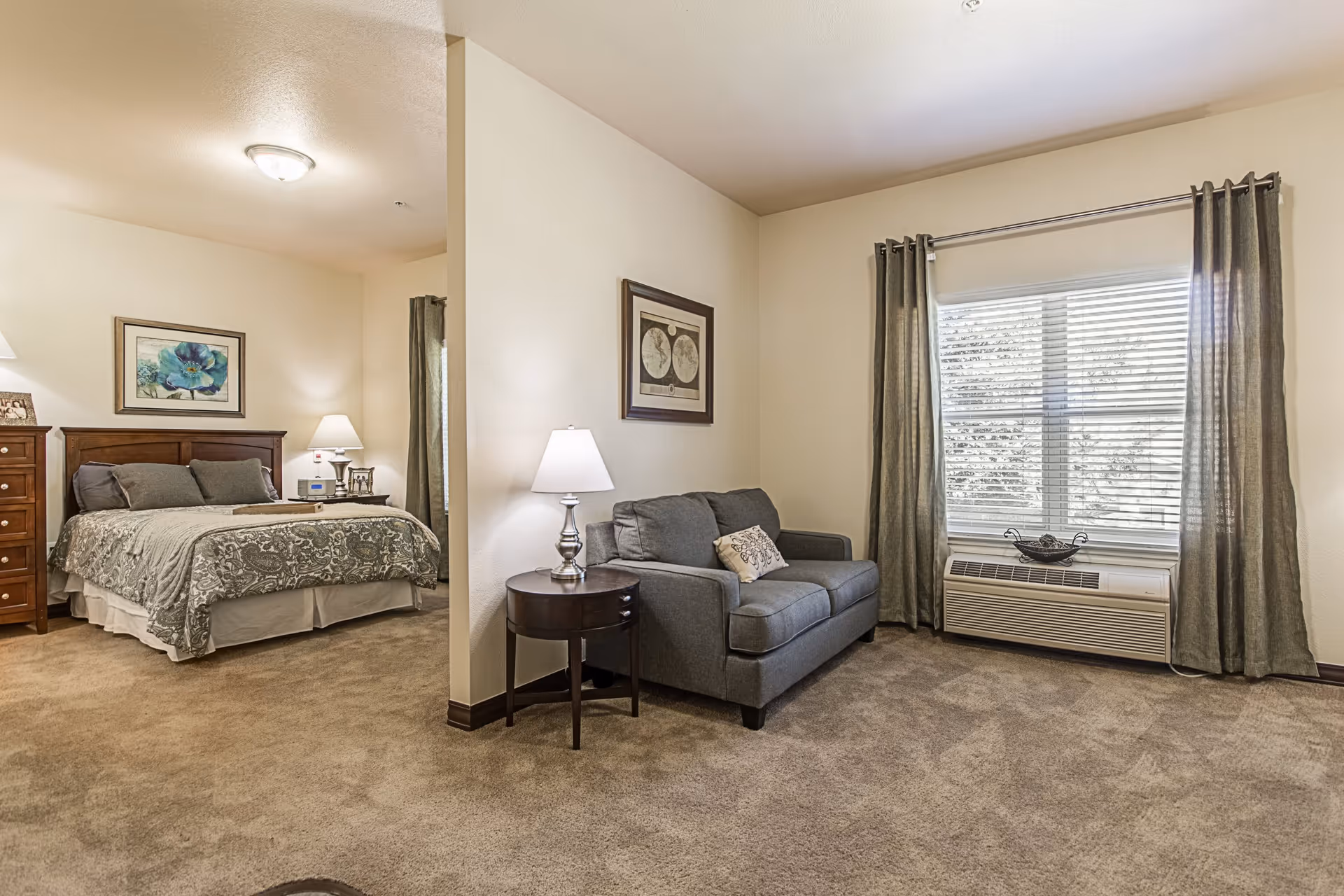 A cozy living space in a senior living facility featuring a small gray loveseat with a decorative pillow, a round side table with a lamp, and a large window with curtains. Adjacent to the living area is a bedroom with a wooden bed frame, a patterned bedspread, two pillows, a nightstand with a lamp, and a dresser. The walls are decorated with framed artwork, and the carpet is beige.