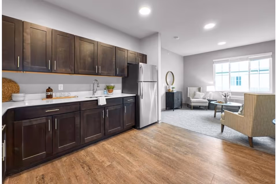 A modern kitchen area with dark wooden cabinets, a white countertop, a stainless steel refrigerator, and a sink. Adjacent to the kitchen is a living room space with two armchairs, a round coffee table, a small black cabinet with a round mirror above it, and a large window letting in natural light.