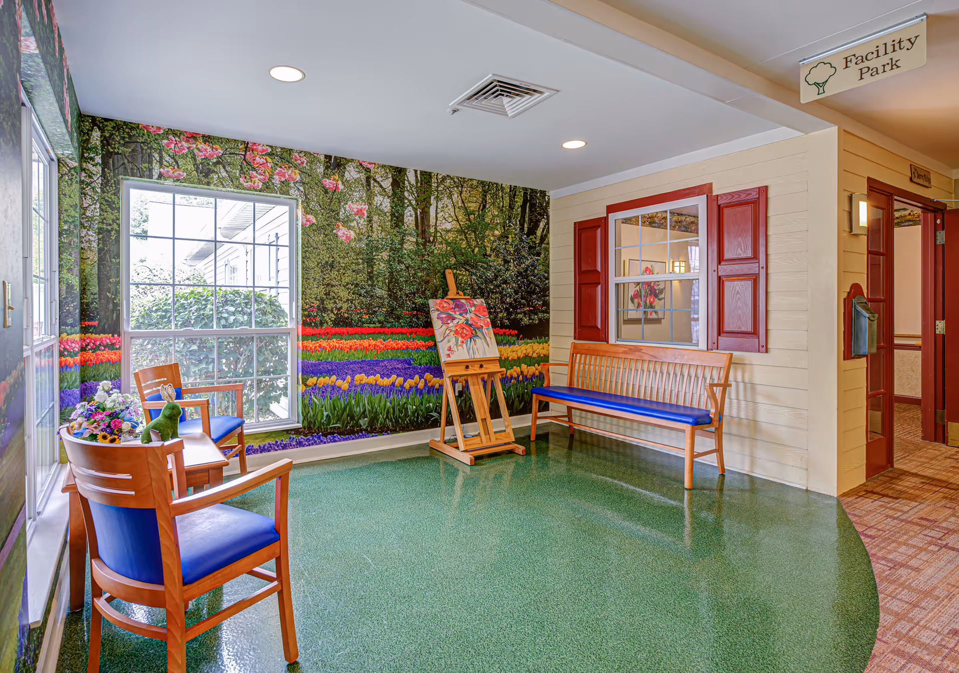 Bright indoor seating area with wooden chairs, a bench, an easel and a large floral mural with a window.