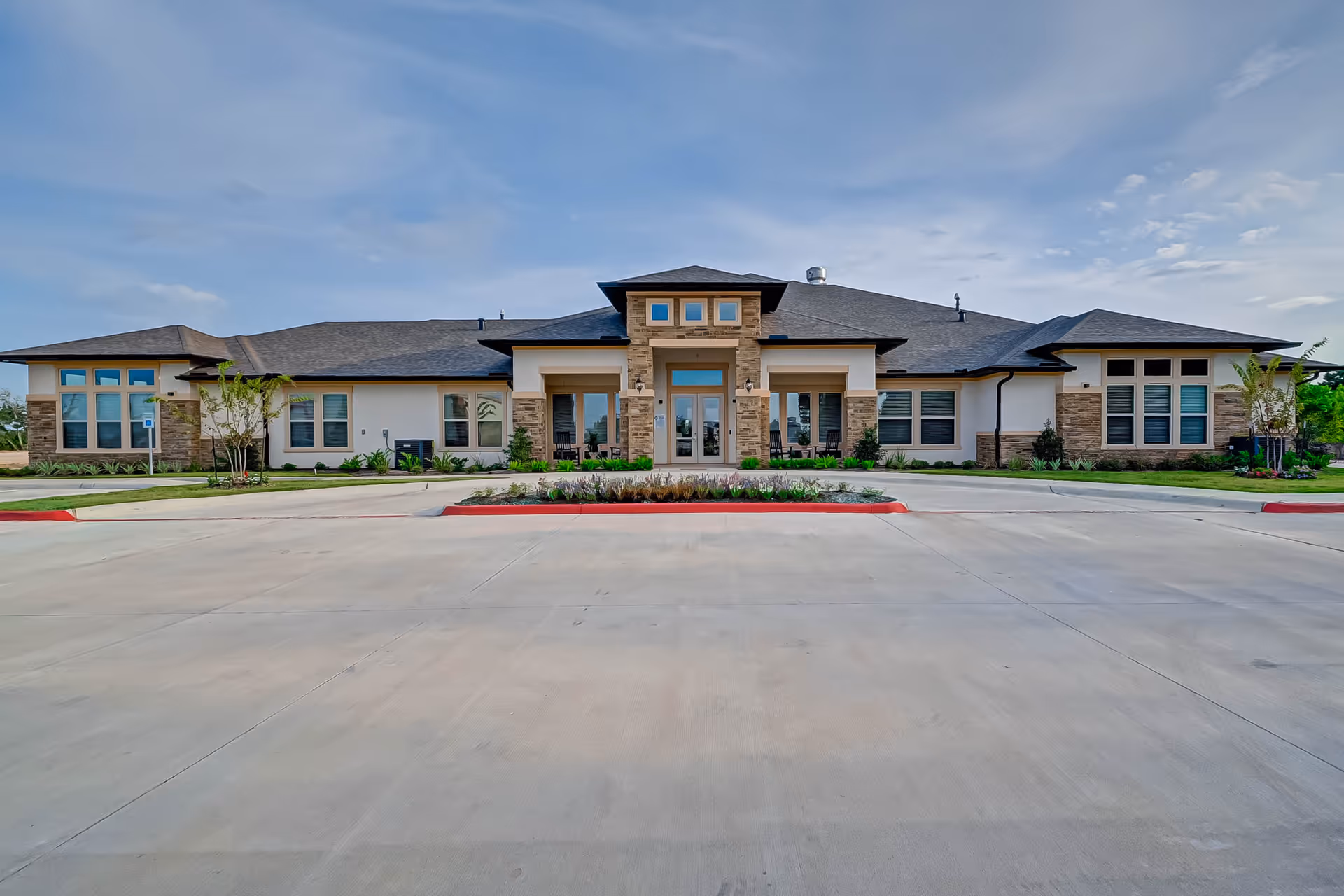 Front exterior view of a single-story assisted living and memory care facility building with a wide driveway and landscaped garden in front under a partly cloudy sky.