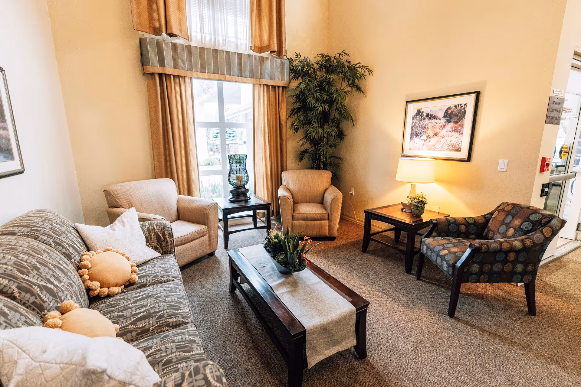 A cozy living room area with a patterned sofa adorned with decorative pillows, two beige armchairs, and a polka-dotted armchair. There is a wooden coffee table with a beige runner and a plant centerpiece. A side table with a lamp and a framed picture on the wall add to the warm ambiance. Large windows with beige curtains allow natural light to fill the room.