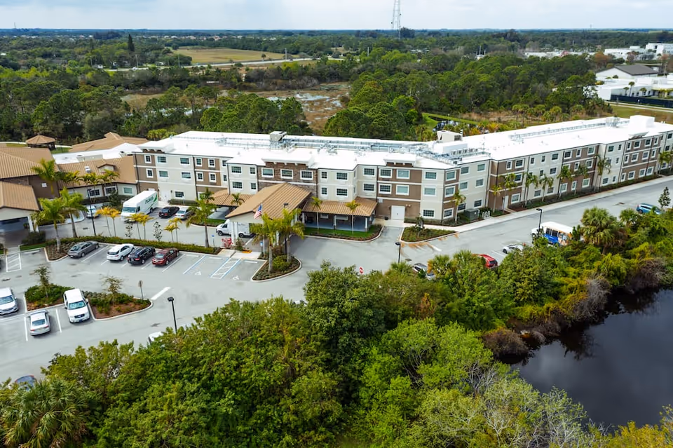 Aerial view of Discovery Village At Stuart, a large senior living facility surrounded by greenery and a small body of water. The building has a beige and white exterior with a brown roof, and there is a parking lot with several cars in front of the facility.