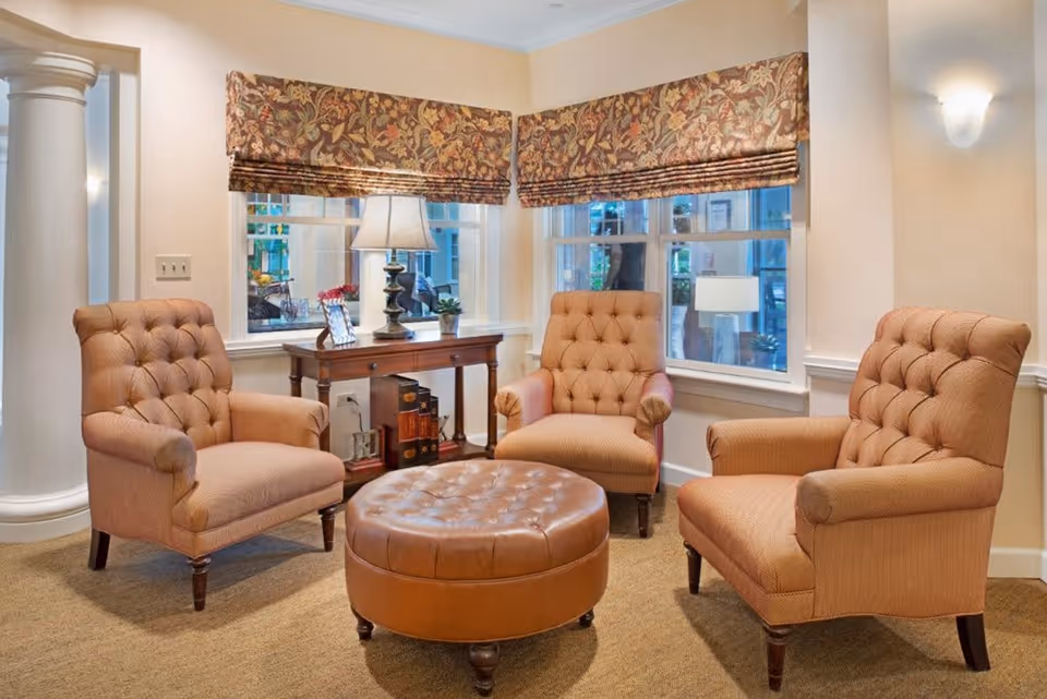 A cozy sitting area with three tufted armchairs arranged around a round, tufted leather ottoman. Behind the chairs is a wooden console table with a lamp, picture frame, and small plant. Two windows with floral patterned valances let in natural light, and the walls are painted a soft beige color.