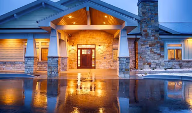 Entrance of a building with stone and wood exterior, illuminated by warm lights at dusk, with a wet reflective driveway in front.
