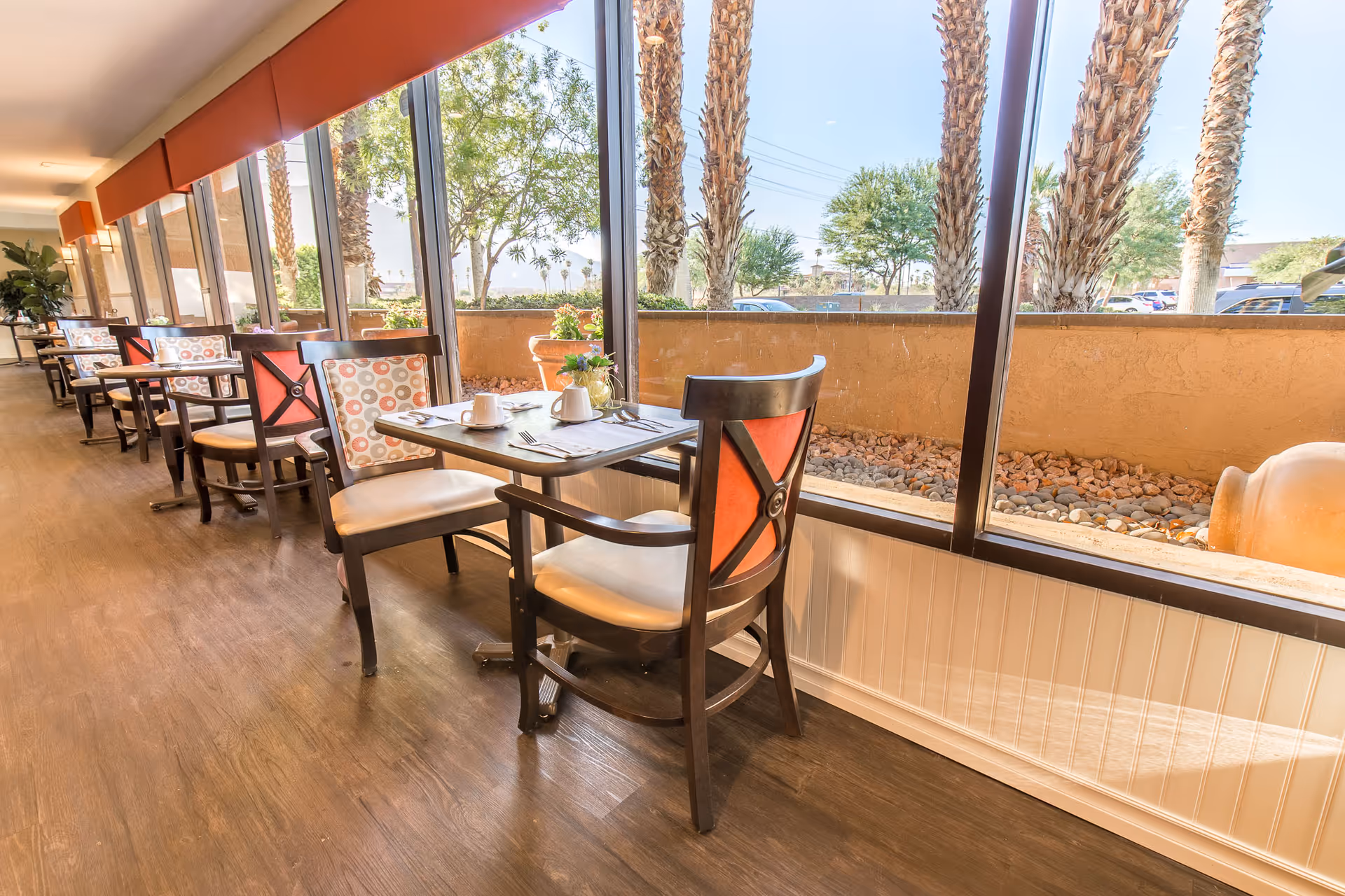A bright dining area with multiple tables and chairs arranged along large windows. The tables are set with cups, plates, and utensils. Outside the windows, palm trees and other greenery are visible under a clear sky.