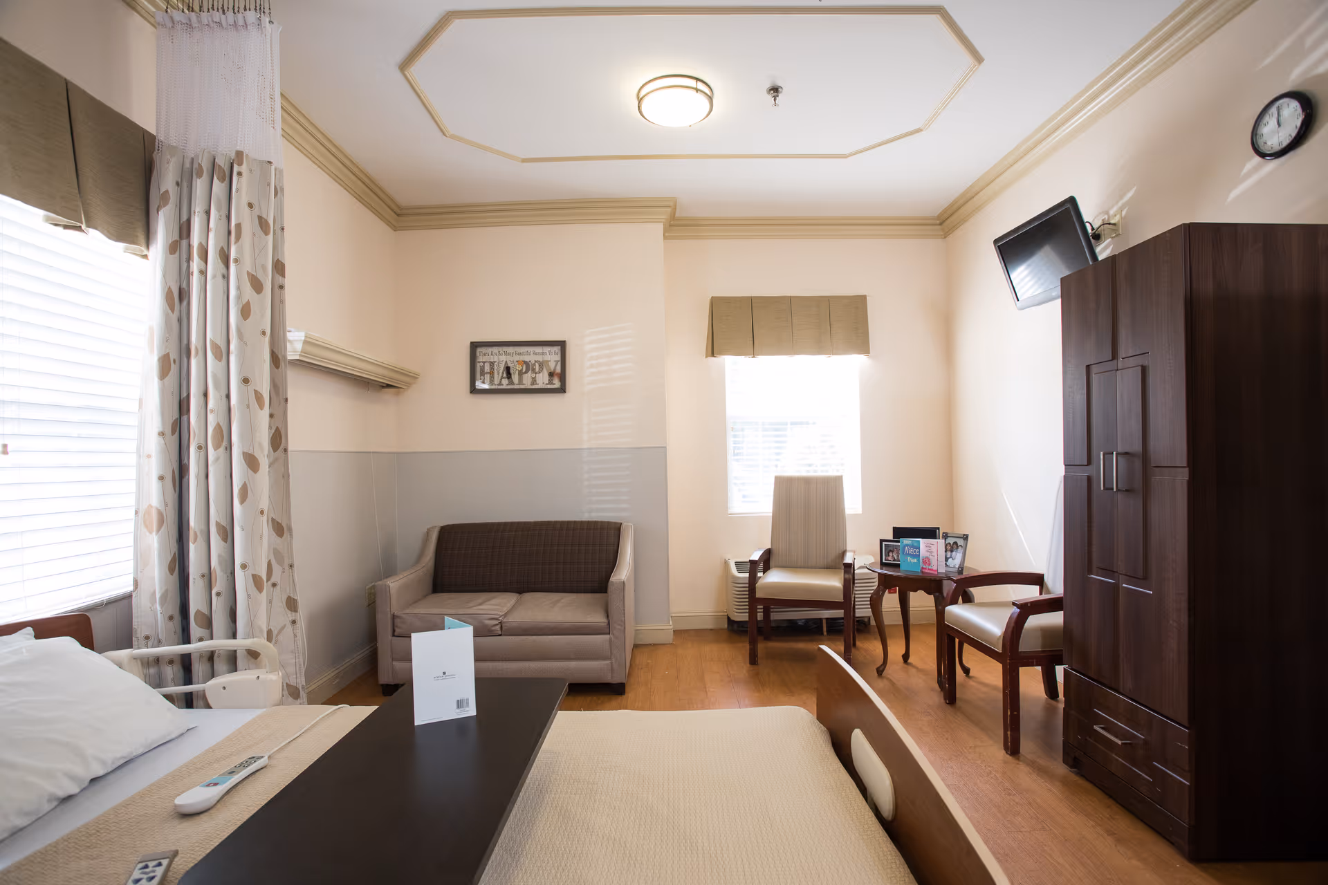 A well-lit room with a hospital-style bed in the foreground, a brown sofa against the wall, two armchairs with a small table between them, a dark wooden wardrobe, and a wall-mounted TV. The room has two windows with blinds and curtains, light-colored walls with crown molding, and a ceiling light fixture.