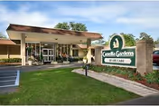 Exterior view of Camellia Gardens of Life Care facility showing the entrance with a covered drop-off area, a well-maintained lawn, and a sign displaying the facility's name near the sidewalk.