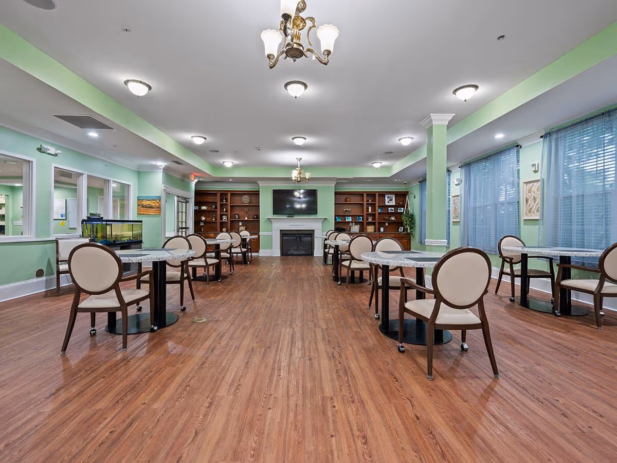 A spacious common area with wooden flooring, light green walls, and multiple tables with beige cushioned chairs arranged neatly. The room features a fireplace with a TV mounted above it, flanked by built-in wooden shelves displaying decorative items. Large windows with sheer blue curtains allow natural light to enter, and ceiling lights along with chandeliers illuminate the space.