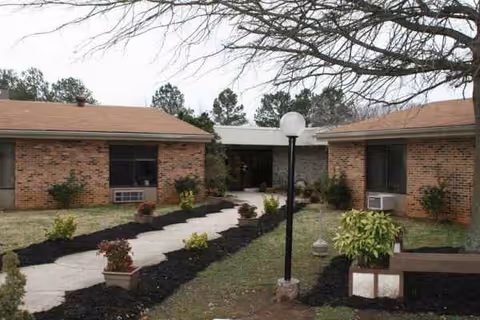 Brick nursing home exterior with central entrance, sidewalk path, and landscaped yard.