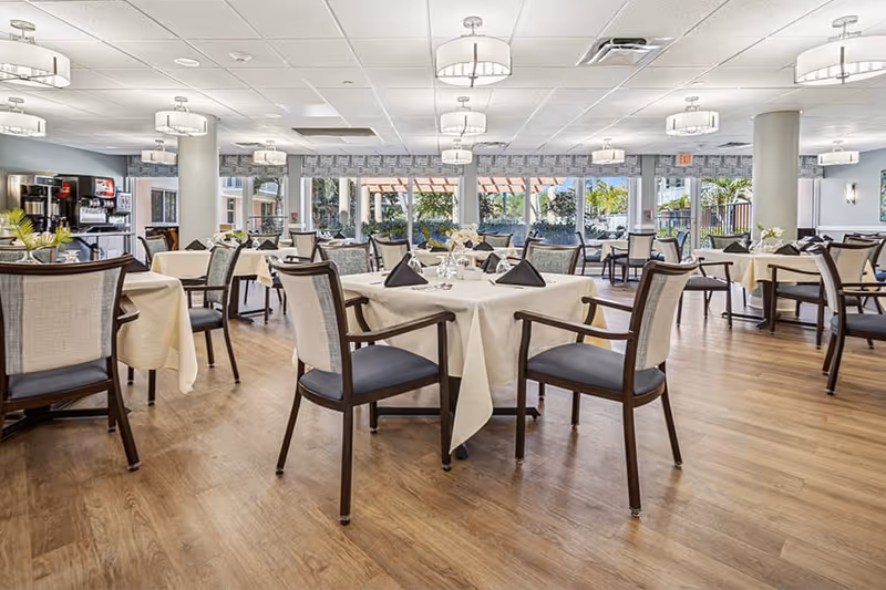 A spacious dining room with multiple tables covered in white tablecloths, each set with black folded napkins and small floral centerpieces. The room has wooden flooring, cushioned chairs with wooden frames, large windows letting in natural light, and modern ceiling lights. In the background, there is a beverage station with coffee machines and a view of an outdoor patio area.
