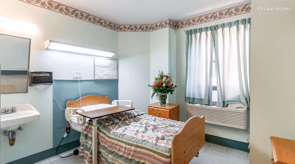 A private room in a healthcare facility featuring a single hospital bed with a striped bedspread, an overbed table, a wooden nightstand with a vase of flowers, a window with light blue curtains, a wall-mounted air conditioning unit, a sink with a mirror above it, and soft lighting. The room has light green walls with a floral border near the ceiling.