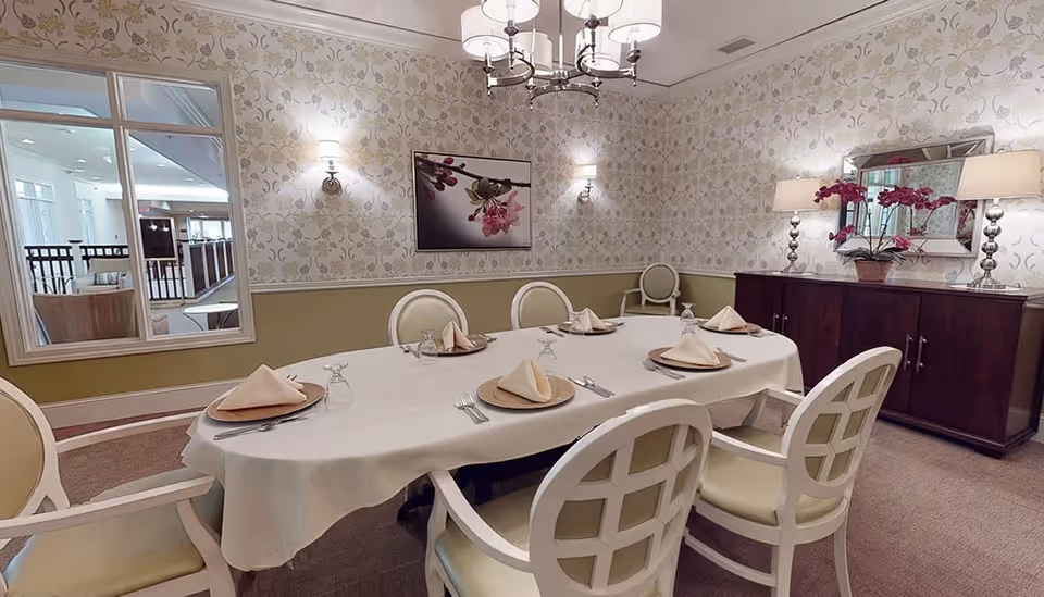 A formal dining room with a long oval table covered with a white tablecloth set for six people. Each place setting has a plate with a folded napkin, a fork, knife, and an upside-down glass. The room has patterned wallpaper, a large mirror above a dark wooden sideboard with two lamps and a flower arrangement. A chandelier hangs above the table, and there is a window looking into another room.