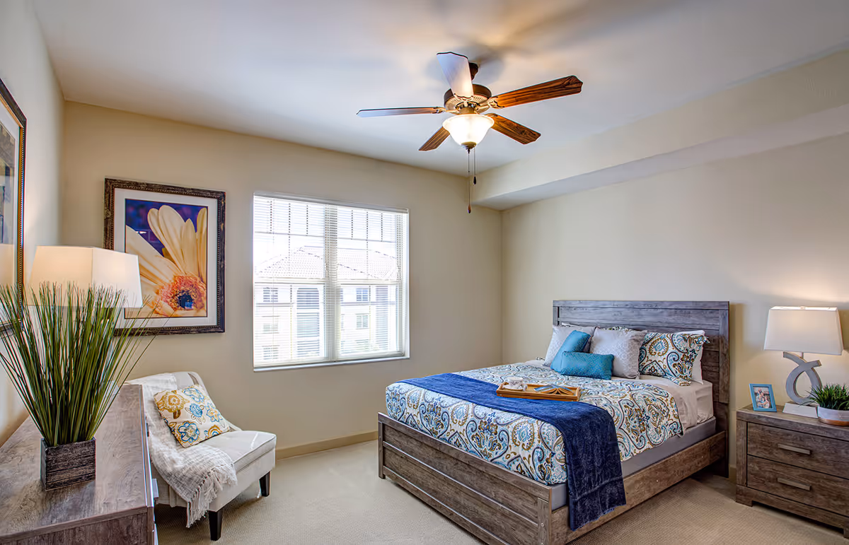 A bright bedroom with a large window letting in natural light. The room features a wooden bed with patterned bedding and blue accent pillows, a wooden nightstand with a lamp and a small framed photo, a wooden dresser with a lamp and a tall plant, and a comfortable chair with a throw blanket and decorative pillow. A ceiling fan with lights is mounted on the ceiling, and a framed flower artwork hangs on the wall.