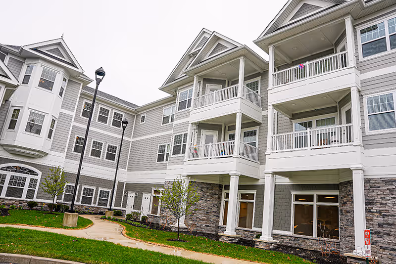Exterior view of a multi-story senior living building with balconies, stone accents, and a landscaped walkway.