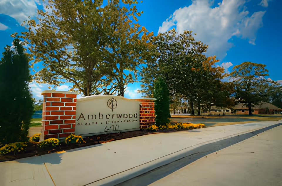 Entrance sign for Amberwood Health + Rehabilitation surrounded by brick pillars, greenery, and flowers with a clear blue sky and trees in the background.