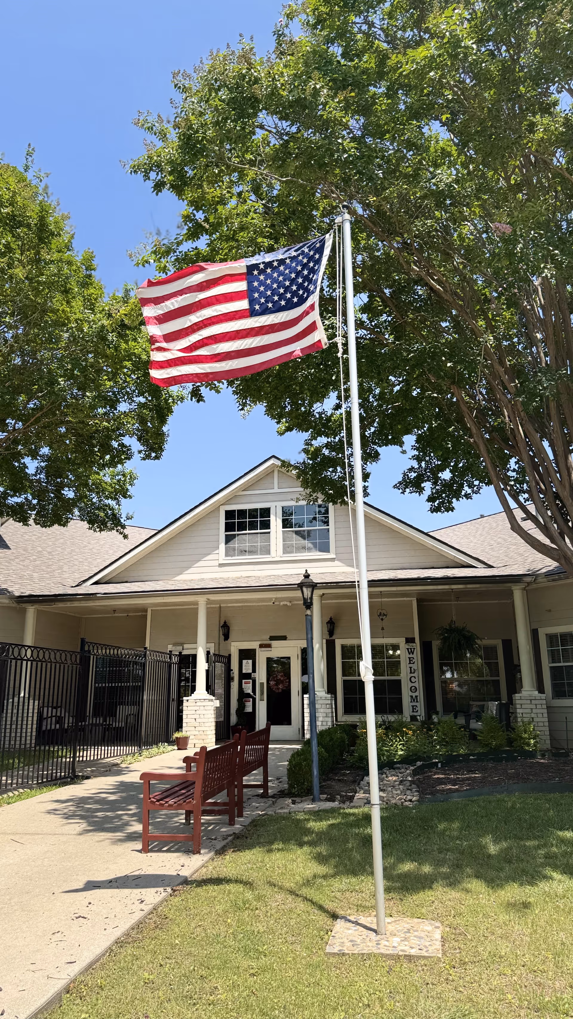 Exterior view of a single-story building with a peaked roof, surrounded by trees and greenery. An American flag is flying on a flagpole in front of the building. There are two red benches on a sidewalk leading to the entrance, which has a 'WELCOME' sign near the door.