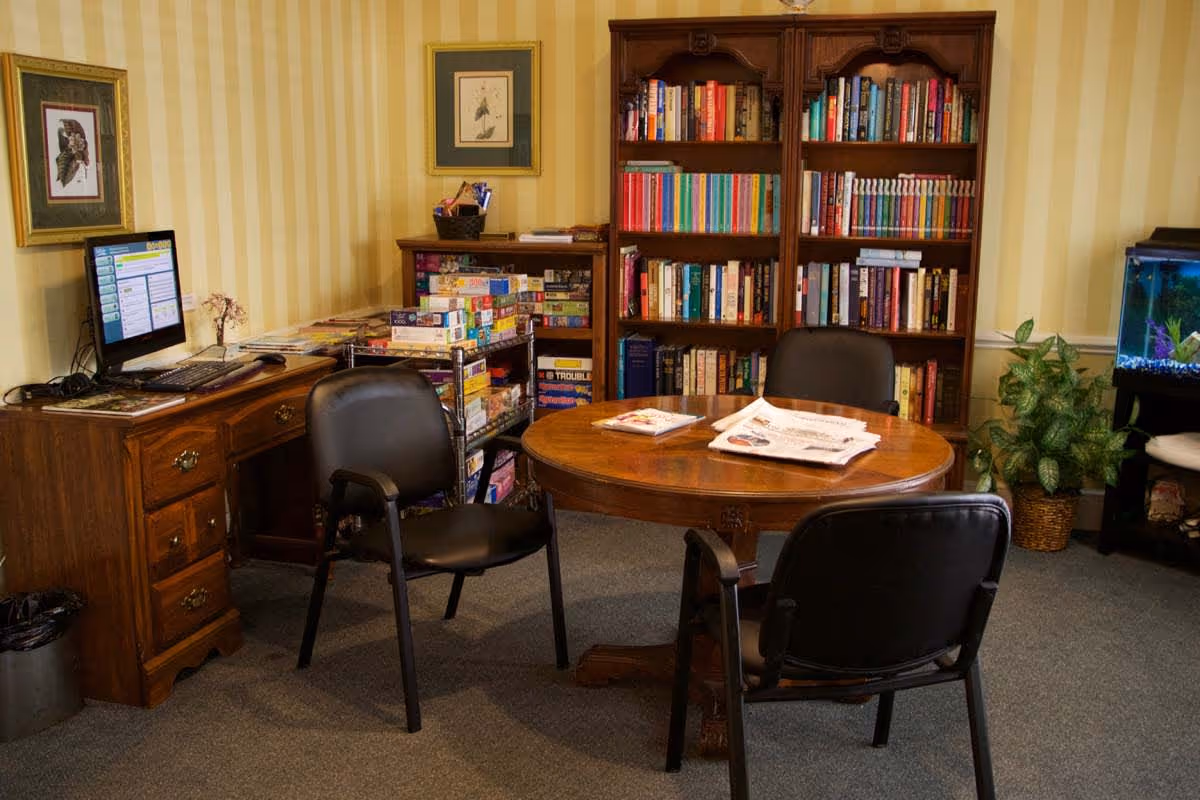 A cozy room with a round wooden table surrounded by three black chairs. Behind the table is a large wooden bookshelf filled with books. To the left, there is a wooden desk with a computer monitor and keyboard, and a metal cart stacked with board games. On the right side, there is a fish tank and a potted plant. The walls have yellow striped wallpaper and framed artwork.