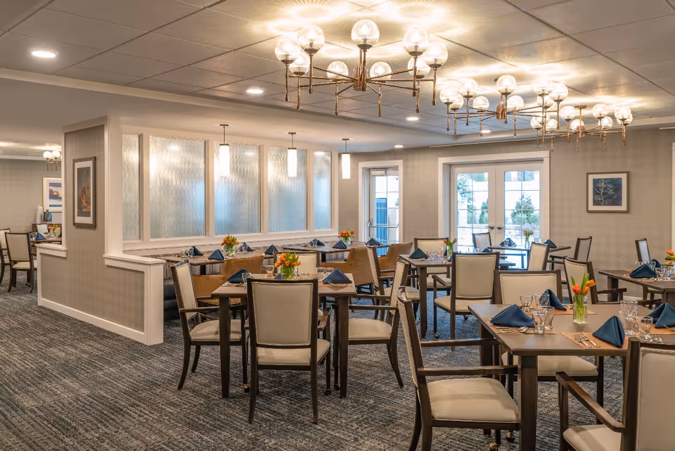 A well-lit dining room with multiple tables set with blue napkins, glassware, and small flower arrangements. The room features modern chandeliers, pendant lights, and large windows with doors leading outside. The decor is neutral with beige walls and carpeted flooring.