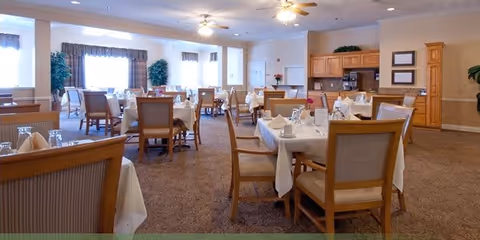 A spacious dining room with multiple tables covered in white tablecloths, set with glassware and napkins. Wooden chairs surround the tables, and the room features carpeted flooring, ceiling fans, large windows with curtains, and a kitchenette area with wooden cabinets in the background.