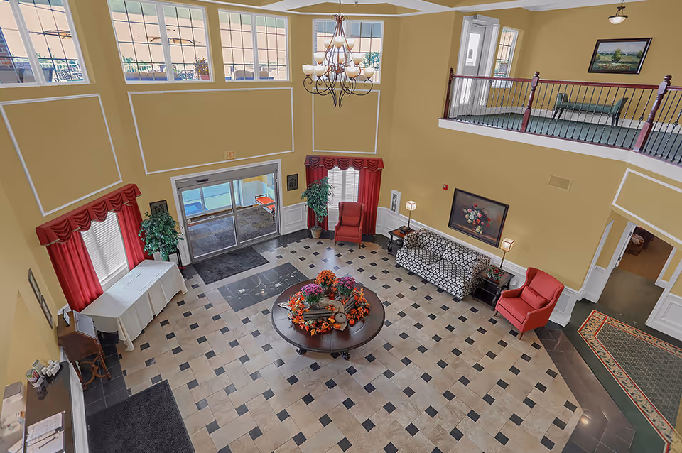 Two-story senior living lobby with a central round table of flowers, seating areas, tiled floor and staircase balcony.